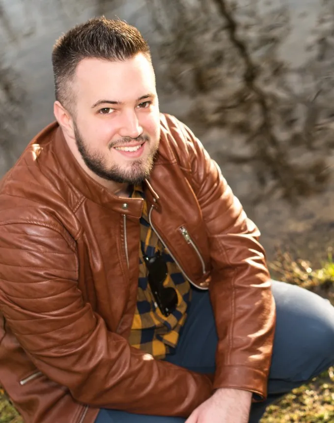 Patrick S. Nussbaumer in brauner Lederjacke, hockend an einem Flussufer, dem Betrachter zugewandt.